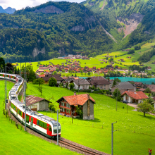 A train travels through a lush green valley with scattered houses, surrounded by towering mountains, under a clear blue sky.