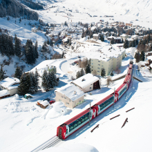 A red train travels through a snow-covered village in the mountains, surrounded by snowy trees and buildings, under a clear sky.