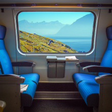 Train interior with blue seats and a scenic window view of rolling hills, vineyards, and mountains by a lake under a clear blue sky.