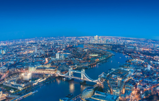 Aerial view of London at dusk, featuring the illuminated Tower Bridge and the winding River Thames under a clear blue sky.