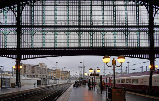 A largely empty train station platform under a high, ornate metal and glass canopy, with a train on the right side and scattered passengers.