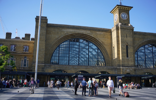 People walking and chatting outside a large historic train station with arched windows and a clock tower under a clear blue sky.