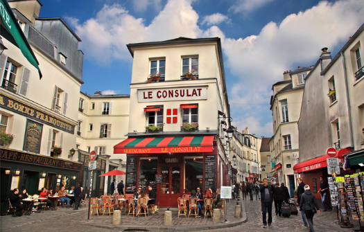 A charming Parisian street scene with the iconic Le Consulat cafe at a corner, surrounded by people, colorful awnings, and historic buildings.