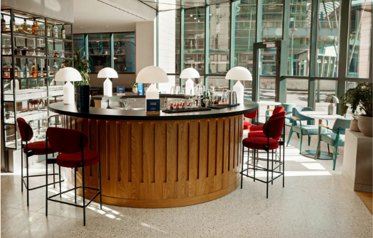 Modern bar area with round wooden counter, red cushioned stools, elegant lamps, and large glass windows letting in natural light.