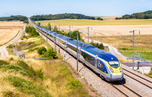 A modern high-speed train travels through a scenic rural landscape, surrounded by grassy fields and distant hills under a clear blue sky.