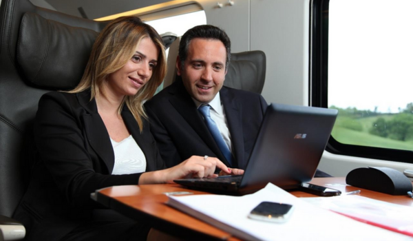 Two professionals collaborate on a laptop while sitting in a train compartment, with papers and a smartphone on the table.