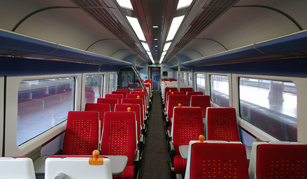 Interior of a train carriage with rows of empty red and white seats, large windows, and overhead storage, viewed from the aisle.