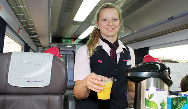 Train attendant in a uniform smiling while serving orange juice in a first-class train carriage.