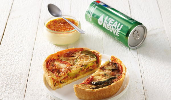 Vegetable quiche slices on a plate with a small jar of dessert and a green soda can on a white wooden table.