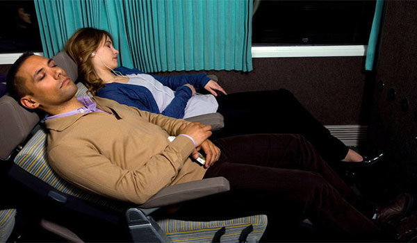 Two people sleeping in reclined seats on a bus or train, with blue curtains in the background.