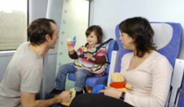 A child sits in a train seat holding a toy, with an adult kneeling nearby and another sitting beside, all smiling and interacting warmly.