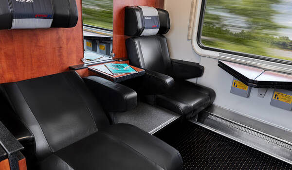 Luxurious train interior with two black leather seats, a fold-out table with a magazine, and a window view of blurred green landscape.
