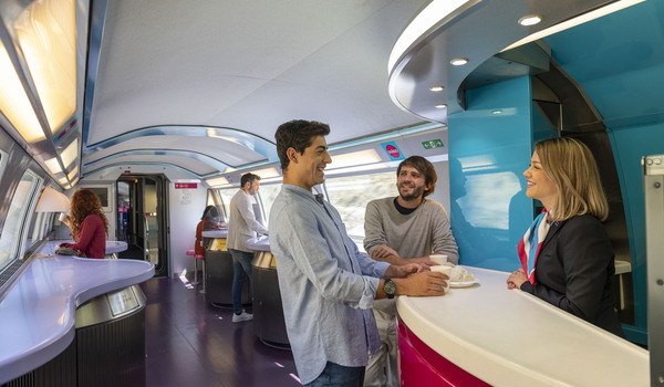 People chatting at a modern train cafe counter, with a curved design and bright lighting, while others are seated further back.