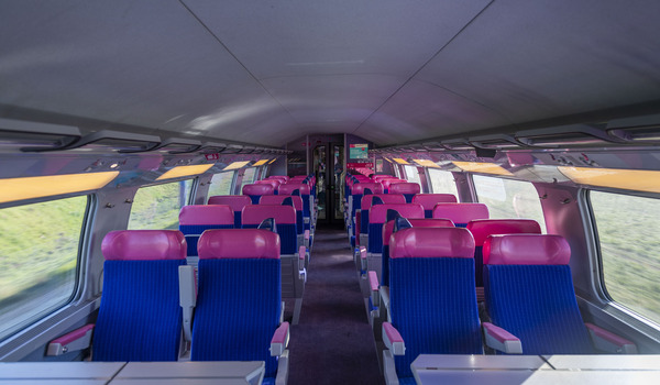Interior of an empty train car with rows of pink and blue seats, large windows, and overhead luggage racks.