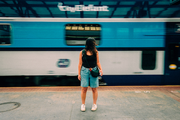 A woman stands on a train platform while a blue train passes by, creating a blurry effect behind her.