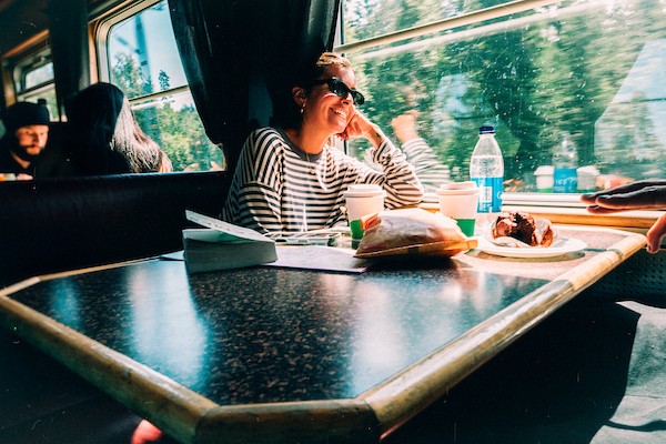 A woman in sunglasses sits at a train table with food and drinks, enjoying the sunlight through the window.