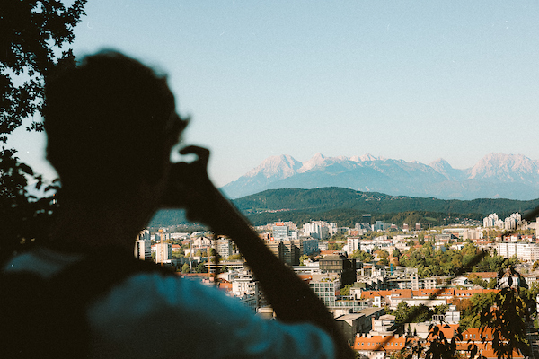 Silhouette of a person photographing a cityscape with distant mountains under a clear blue sky.