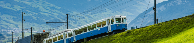 Blue and white train on a steep grassy incline in a mountainous landscape, with overhead power lines and distant hills in the background.