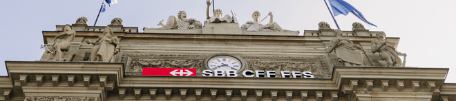 Facade of a historical building with statues, a clock, and the SBB CFF FFS logo, adorned with flags under a clear sky.