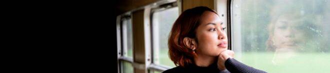 Woman gazes out a train window with her chin resting on her hand, reflecting on the glass, while wearing a black turtleneck and red earrings.