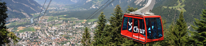 Red cable car over lush pine trees, with a panoramic view of a picturesque valley and distant mountains.