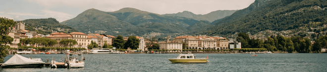 Scenic view of a lake with boats, surrounded by buildings and lush mountains under a cloudy sky.