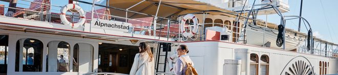 Two people walking towards a docked boat named "Alpnachstad" with lifebuoys, against a clear blue sky.