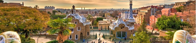 Panoramic view of Park Güell in Barcelona, with Gaudí’s colorful architecture, palm trees, and a vibrant cityscape under a golden sunset sky.