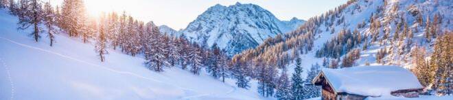 Snow-covered mountains with pine trees, a wooden cabin, and the sun setting in the backdrop, creating a serene winter landscape.