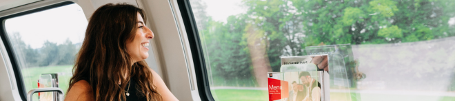 A woman with long hair smiles while looking out a train window at greenery. A brochure holder is visible next to her.