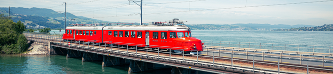 A red train travels over a bridge spanning a large body of water, with distant hills and a partly cloudy sky in the background.
