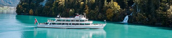 A white ferry with passengers sails on a turquoise lake, surrounded by forested mountains and a small waterfall in the background.