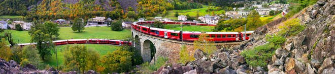 Red train crossing a stone viaduct in a picturesque valley with green fields, trees, and a small village in the background.