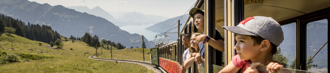 People enjoying mountain views from a train window, with lush green scenery, distant mountains, and a lake in the background.
