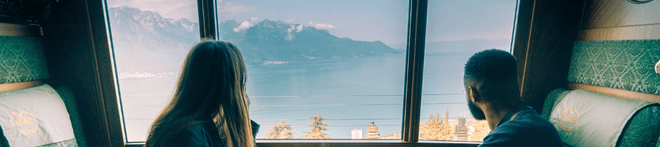 Two people sitting in a train, gazing out at a picturesque lake and mountain landscape through large windows.