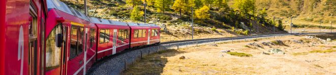 Red train traveling through a scenic mountainous landscape with yellow autumn trees and a clear blue sky.