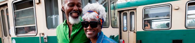 Smiling couple standing in front of a green and beige tram; the man is in a green shirt and the woman wears sunglasses and a colorful headwrap.