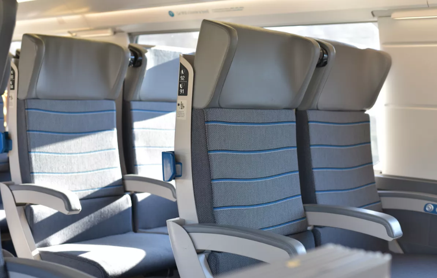 Empty train seats with gray upholstery and blue stripes, featuring armrests and window seats in a well-lit cabin.