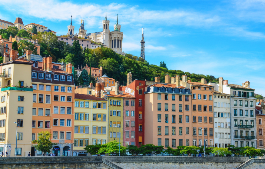 Colorful buildings line a riverbank with a hillside and a basilica in the background under a clear blue sky.